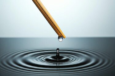 A hand attempts to capture a single droplet of water with a pair of chopsticks, above concentric water ripples.